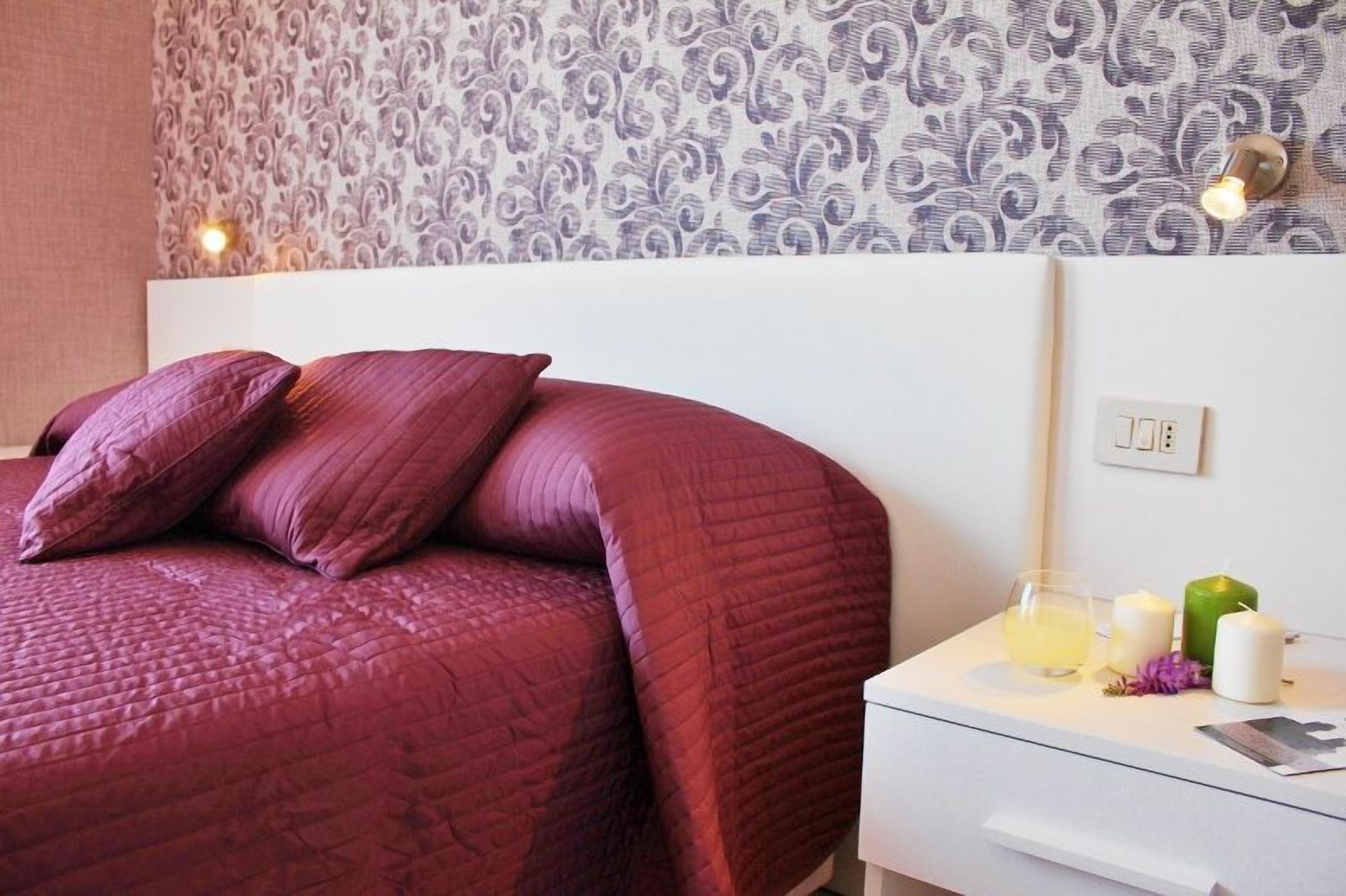 Een net opgemaakt bed met een rode quilt, een witte nachtkastje met een glas water, een kaars en een groene kaars.