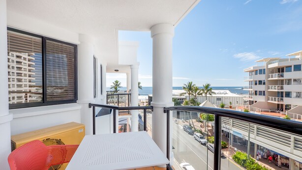 Un balcon avec vue sur la plage et l’océan, une table blanche, une chaise rouge et une fenêtre avec des stores.