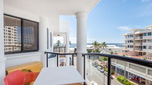 A balcony with a view of the beach and ocean, a white table, a red chair, and a window with blinds. - Aegean Apartments (Mooloolaba)