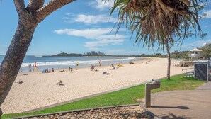 Une plage où l’on peut voir des gens se prélasser au soleil et nager, une tour de surveillance des sauveteurs, et, au loin, la silhouette d’une ville.