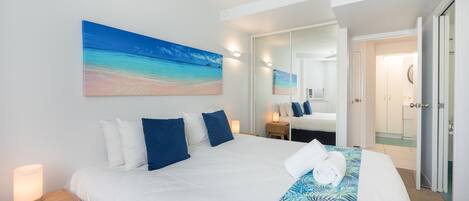 Una habitación de hotel con una cama grande, una mesita de noche, una lámpara de noche, un espejo de baño y una puerta que da a otra habitación.