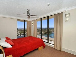 A hotel room with a large bed, a ceiling fan, and a view of a cityscape through large windows. - Alexander Holiday Apartments (Surfers Paradise)