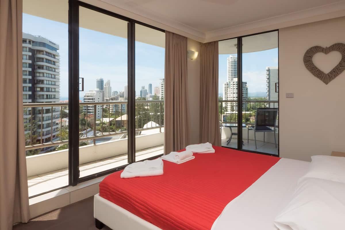 Une chambre d’hôtel avec un grand lit, une vue sur la ville grâce à de grandes fenêtres du sol au plafond, et une décoration en forme de cœur sur le mur.