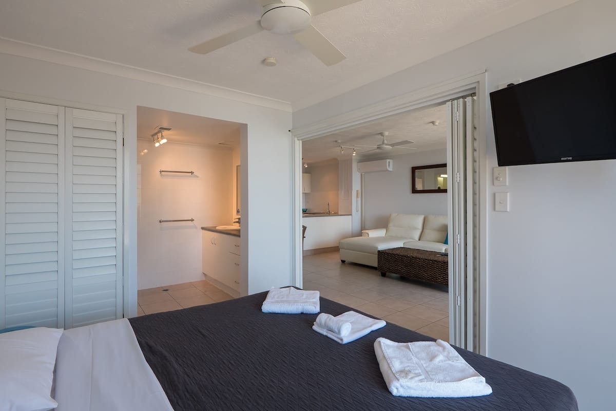 Une chambre à coucher avec un lit, deux serviettes, un ventilateur de plafond, une télévision et une salle de bain visible à travers une porte ouverte.
