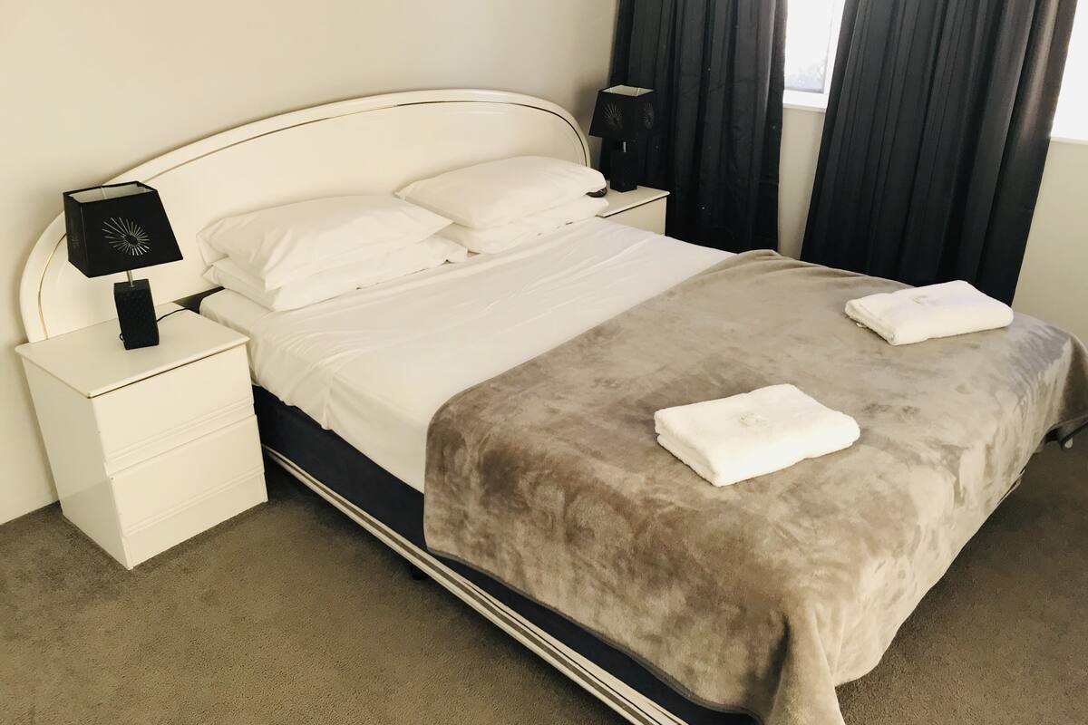 A neatly made bed with a beige duvet and two folded towels, a white bedside table with a lamp, and a window with dark curtains.
