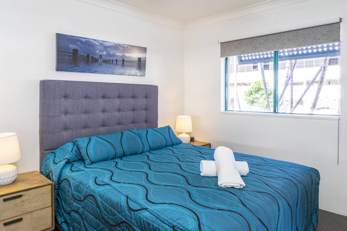 Habitación de hotel con una cama grande, una mesita de noche con lámpara, un cuadro de un atardecer sobre el agua, y una ventana con vista a edificios y árboles.