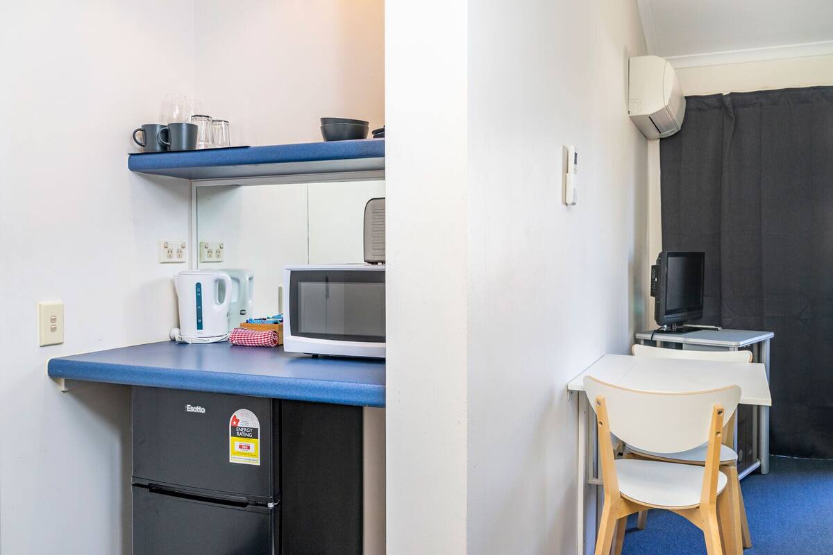 Una kitchenette compacta con microondas, hervidor de agua y un refrigerador pequeño. Hay un televisor y una silla en una zona separada.