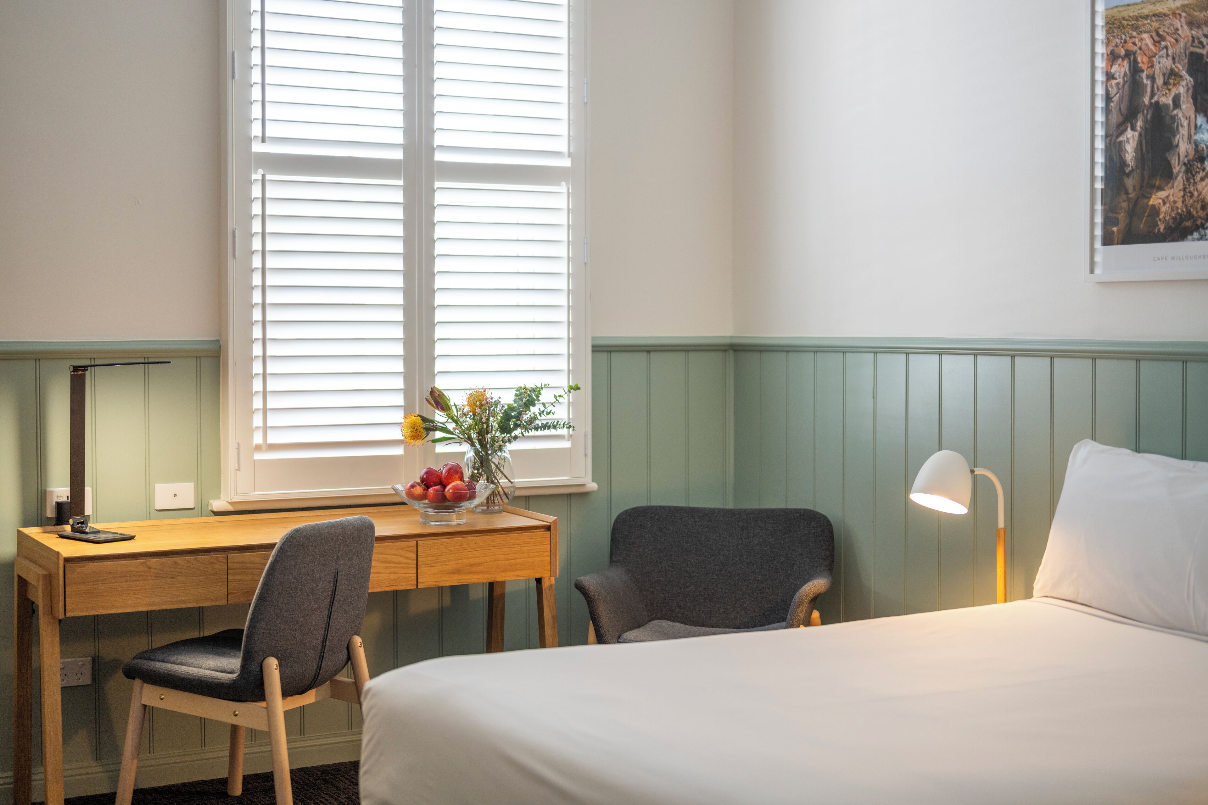 A hotel room with a bed, a desk with a chair, a lamp, and a window with blinds.
