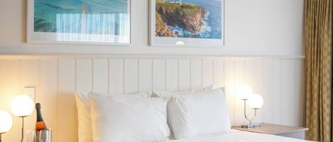 Une chambre d’hôtel avec un lit, deux tableaux représentant des paysages côtiers, une bouteille de champagne et deux lampes de chevet.