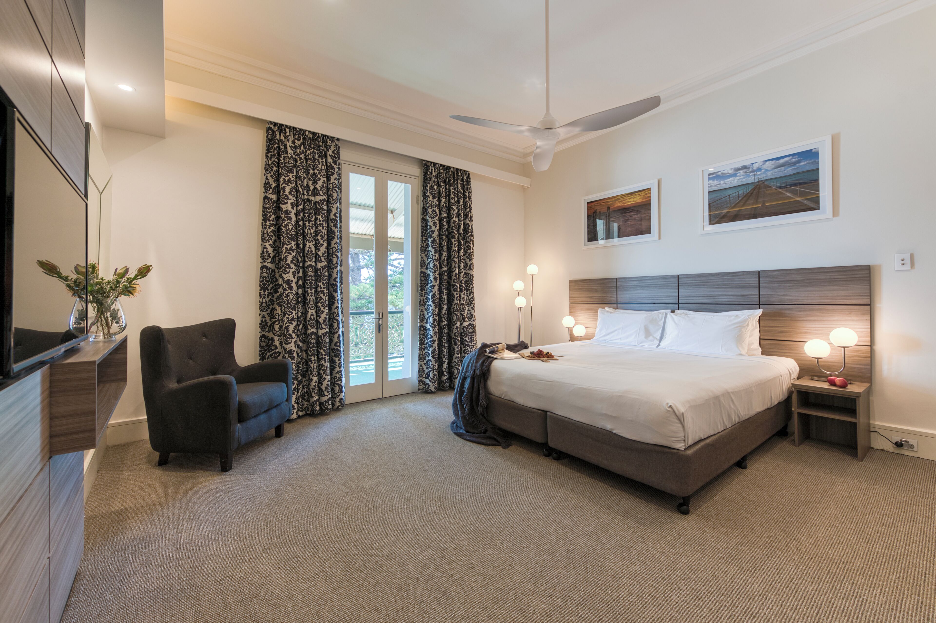 A hotel room with a bed, a chair, a desk, a TV, and a balcony with curtains.