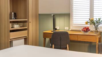 A compact room with a wooden desk, a chair, a small shelf with a kettle, and a window with blinds.