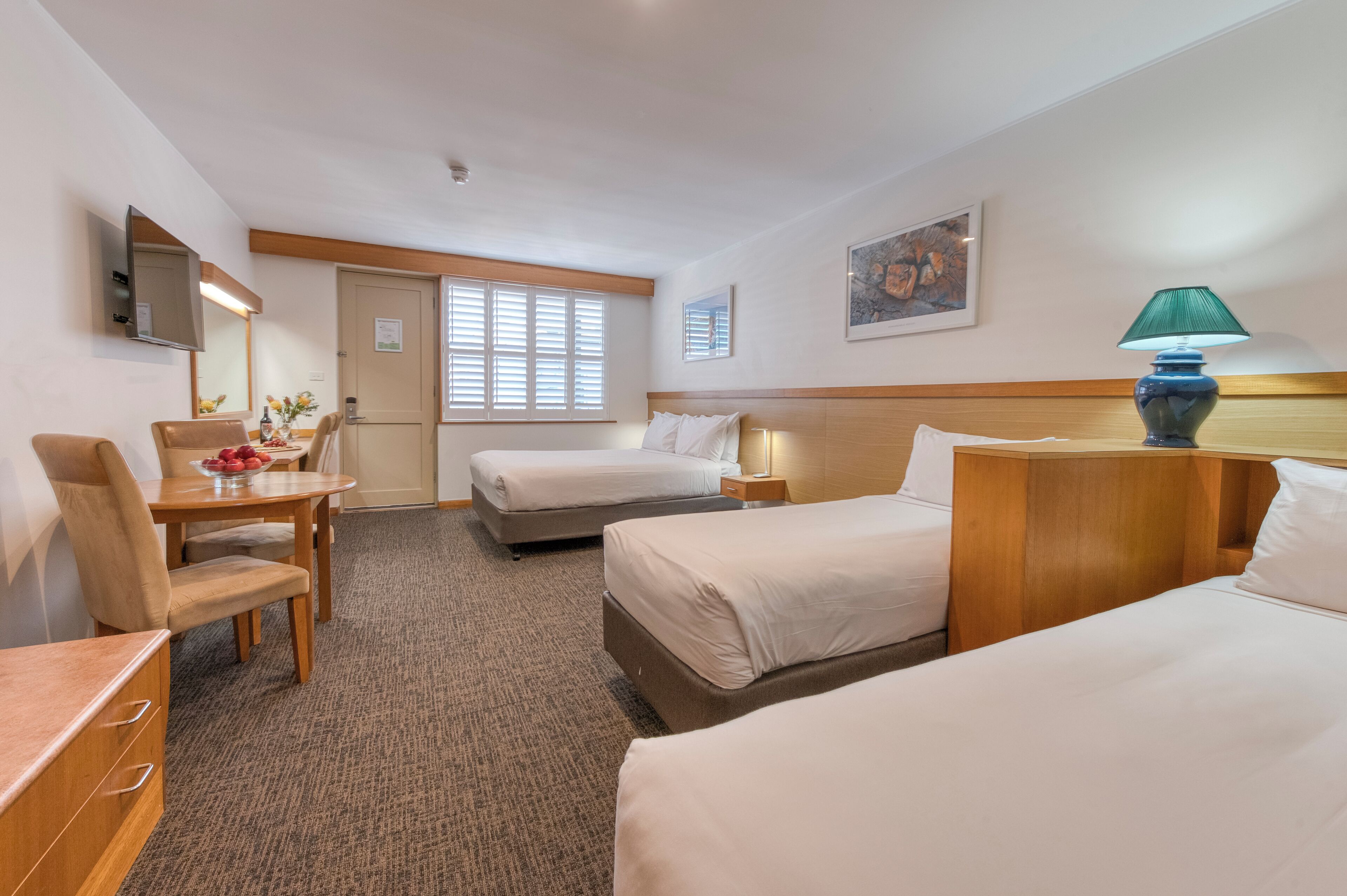 A hotel room with two beds, a desk, a chair, a small table with a vase of flowers, and a lamp.