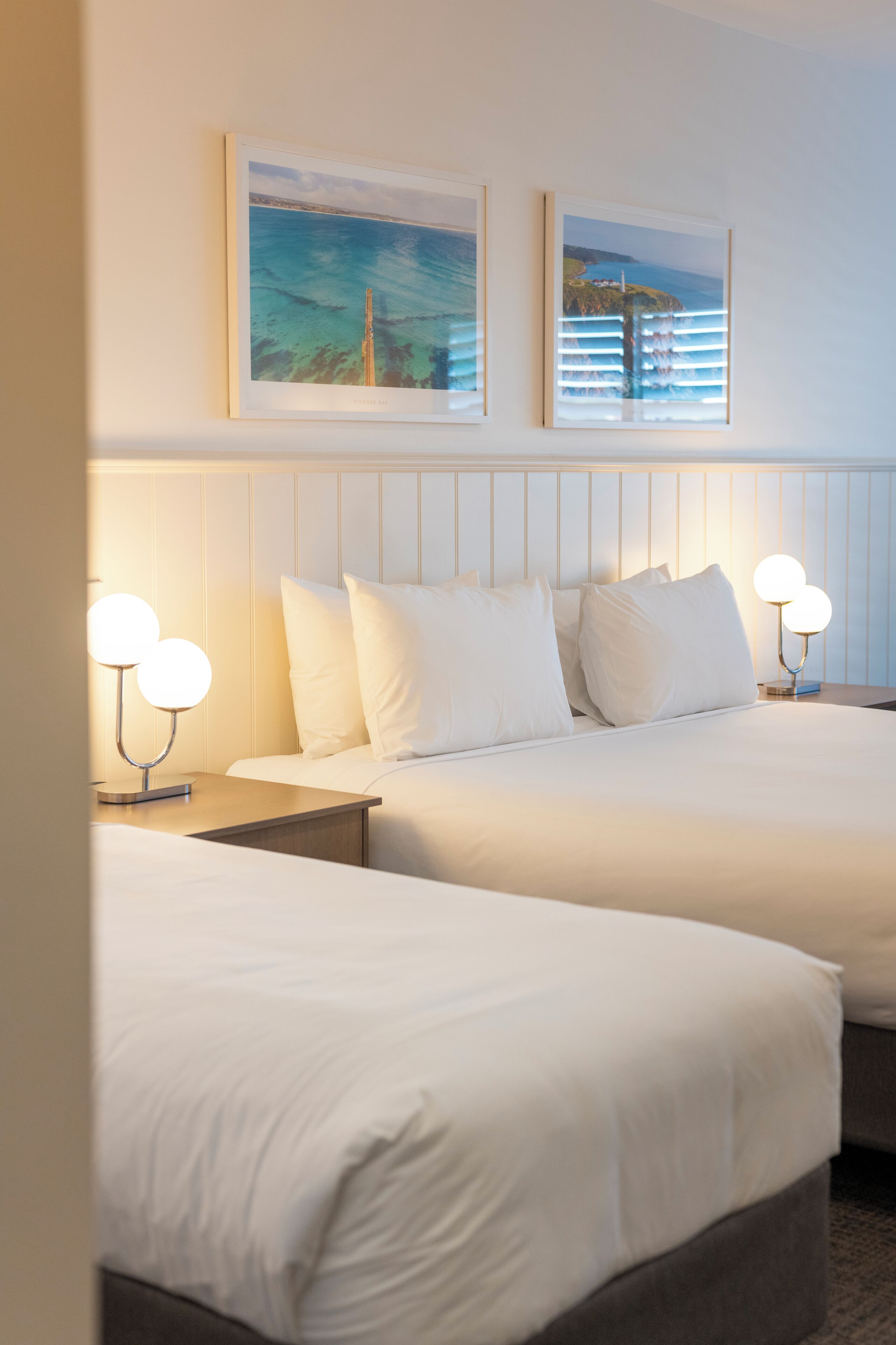 A hotel room with two beds, white bedding, a headboard, bedside tables with lamps, and two framed pictures on the wall.