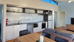 A modern kitchen with white cabinets, a dishwasher, and a built-in oven. There is a dining area with a table and chairs, and a grey sofa. - Serenity Diamond Beach (Diamond Beach)