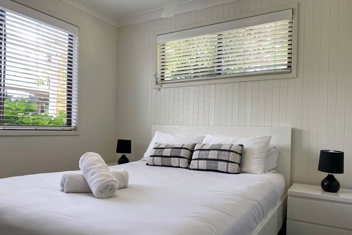A neatly made bed with white linens and a rolled towel, two bedside tables with lamps, and a window with blinds.