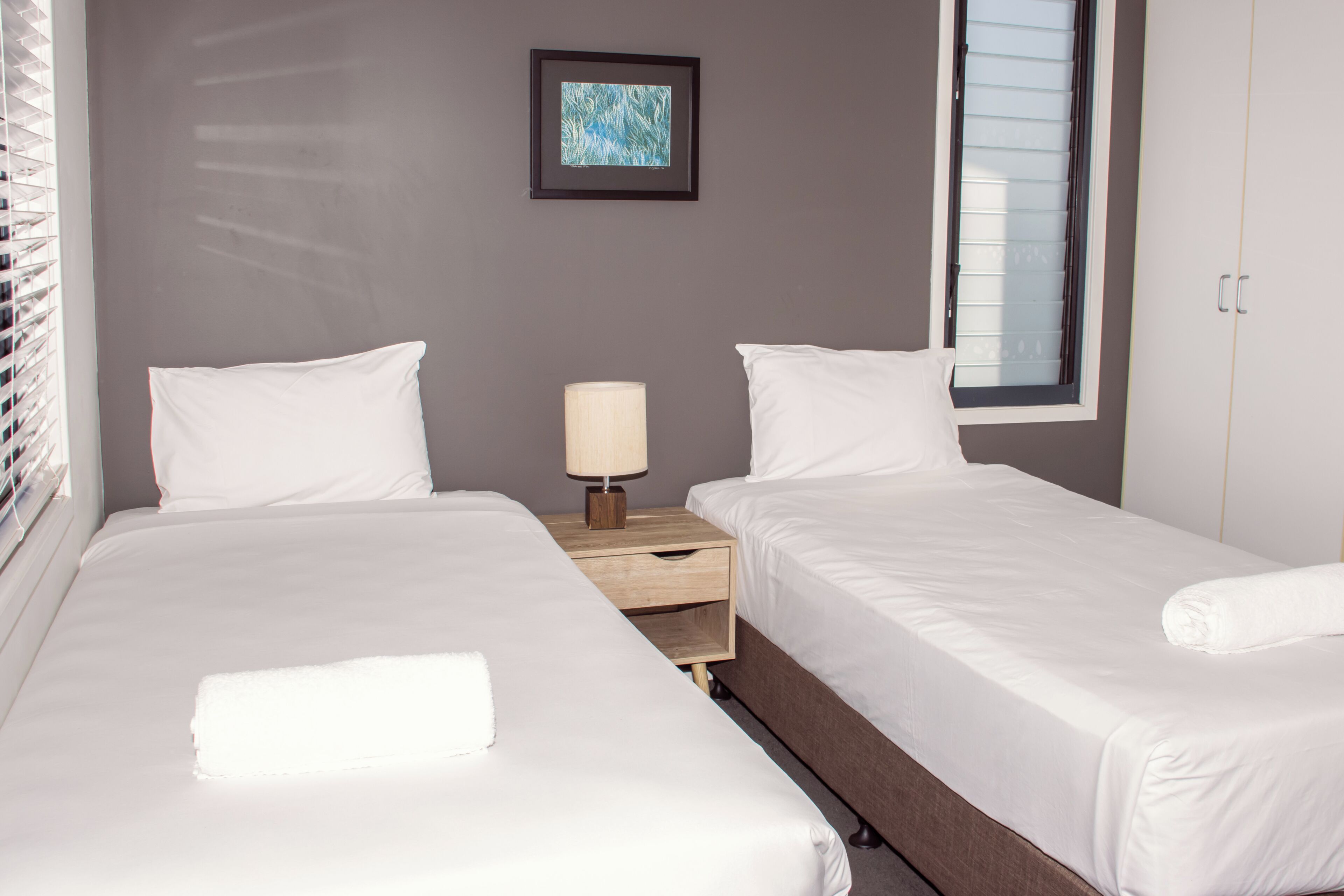 A hotel room with two beds, a nightstand with a lamp, a framed picture on the wall, and a window with blinds.