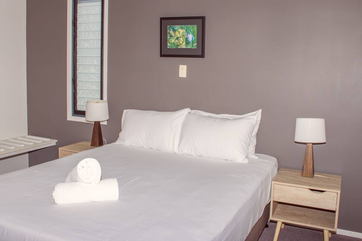 A neatly made bed with white linens and a rolled towel, two bedside tables with lamps, a framed picture on the wall, and a window with blinds.