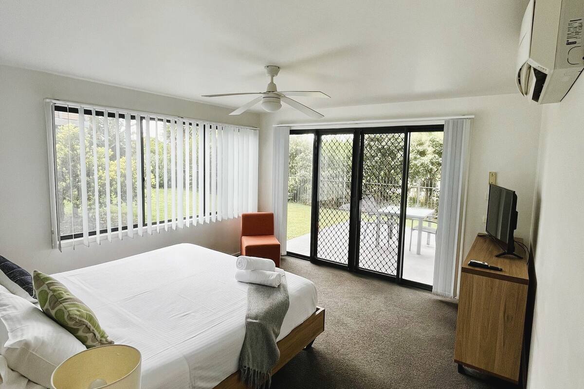 A hotel room with a bed, a desk with a lamp, a TV on a wooden cabinet, and a ceiling fan.