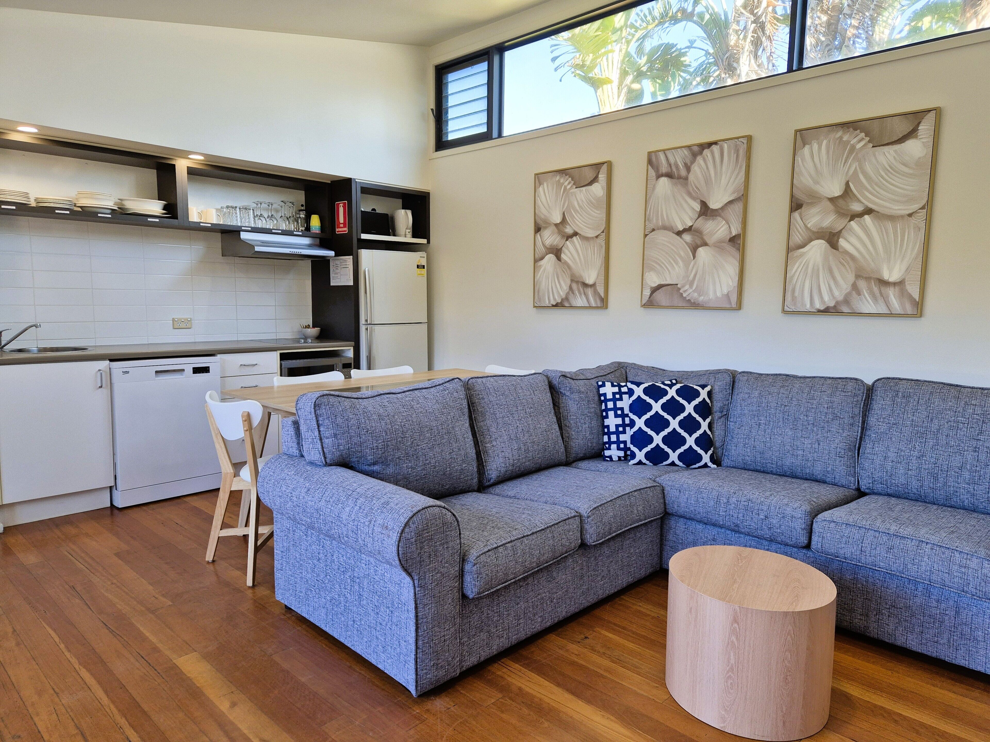 Una sala de estar moderna con un sofá seccional gris, una mesa de centro de madera y una zona de cocina con electrodomésticos y estantes blancos.