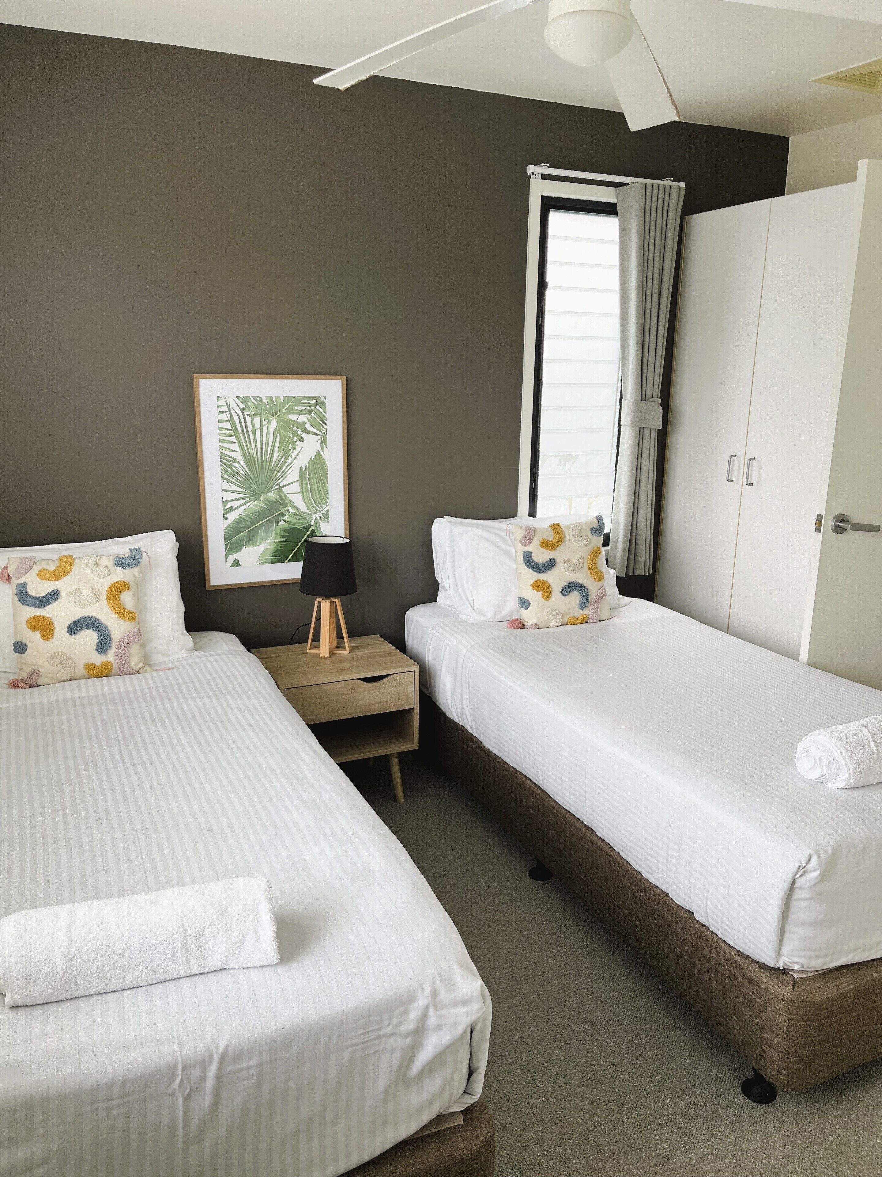 A twin room with two beds, a nightstand, a framed plant, and a wardrobe.
