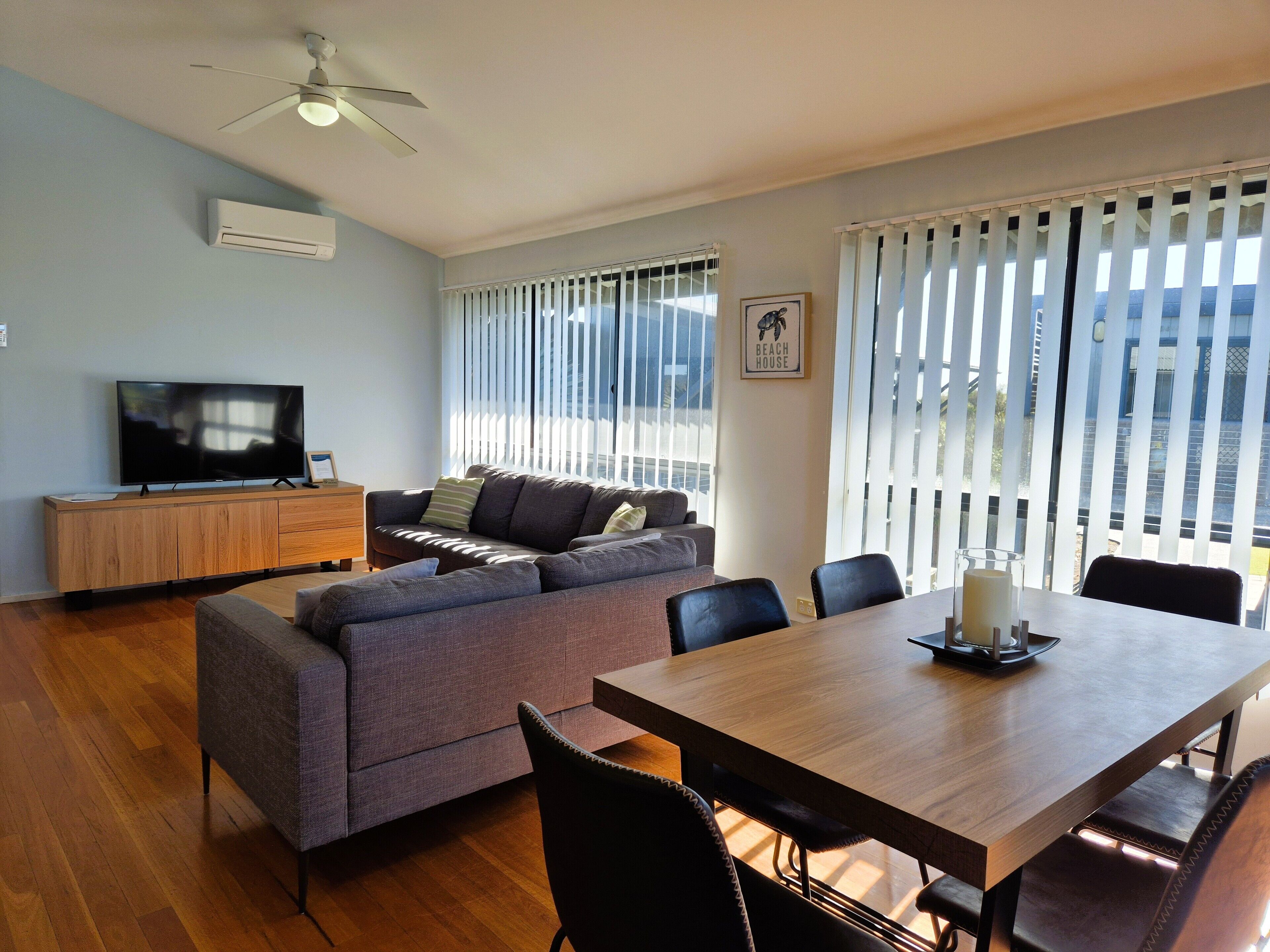 Una sala moderna con sofá, mesa de comedor y sillas. Hay un televisor sobre un mueble de madera, y un ventilador de techo.