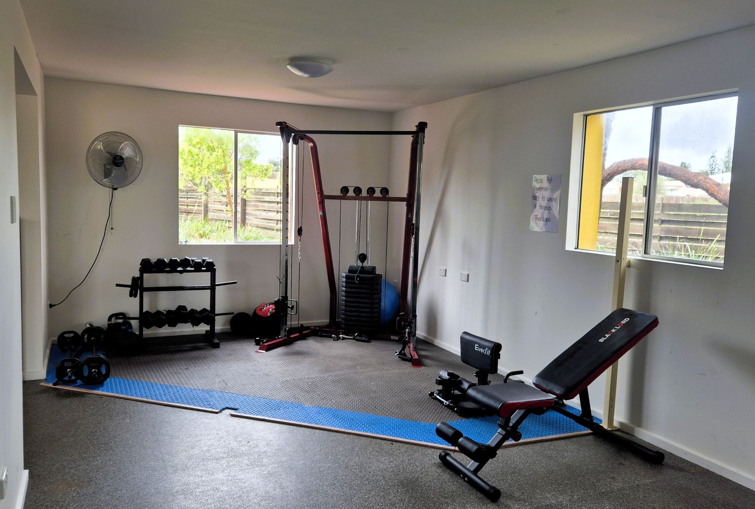Een huisgym met een pull-up bar, een gewichtrek, dumbbells, een bankje en een ventilator.