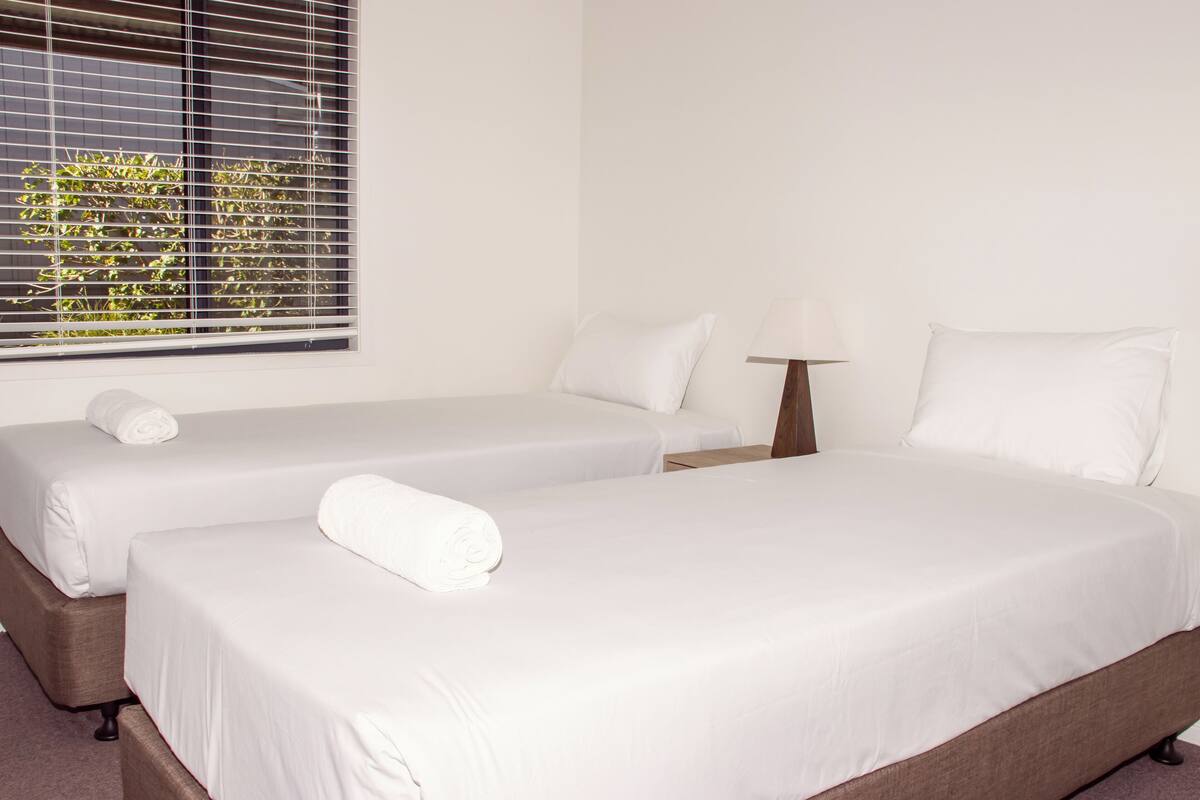 A hotel room with two single beds, a bedside table with a lamp, and a window with blinds.