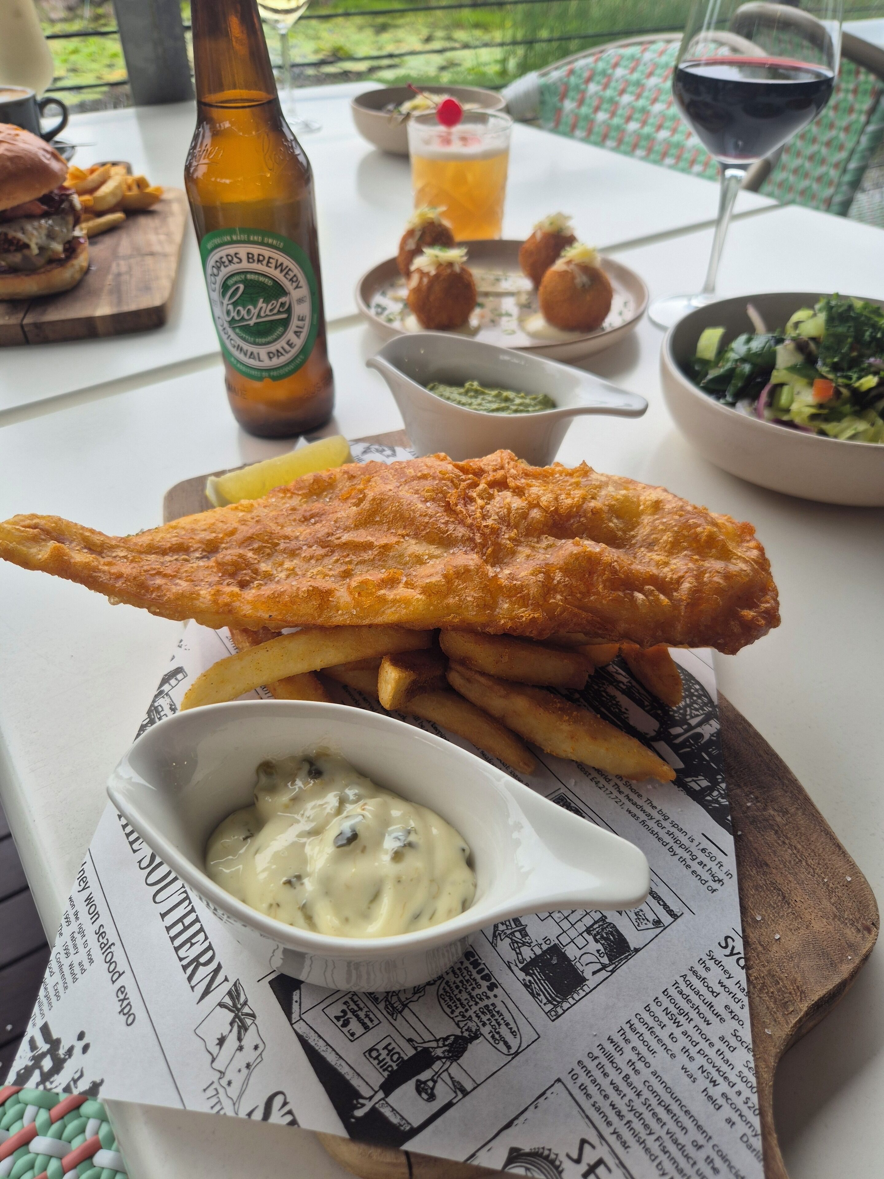 Un plato de pescado con patatas fritas acompañado de salsa, una botella de cerveza, un vaso de cerveza y una ensalada sobre una mesa.