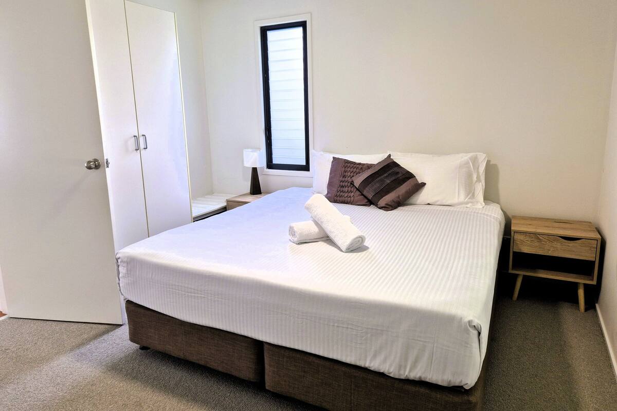 A neatly made bed with a white comforter and two pillows, a wooden nightstand with a lamp, and a white wardrobe with a door.