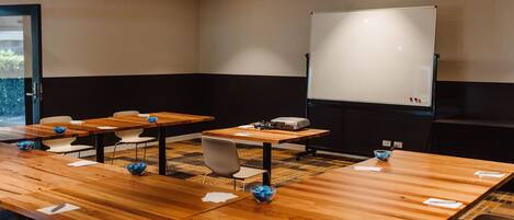 Een vergaderzaal met houten tafels, witte stoelen, een whiteboard en een beamer.