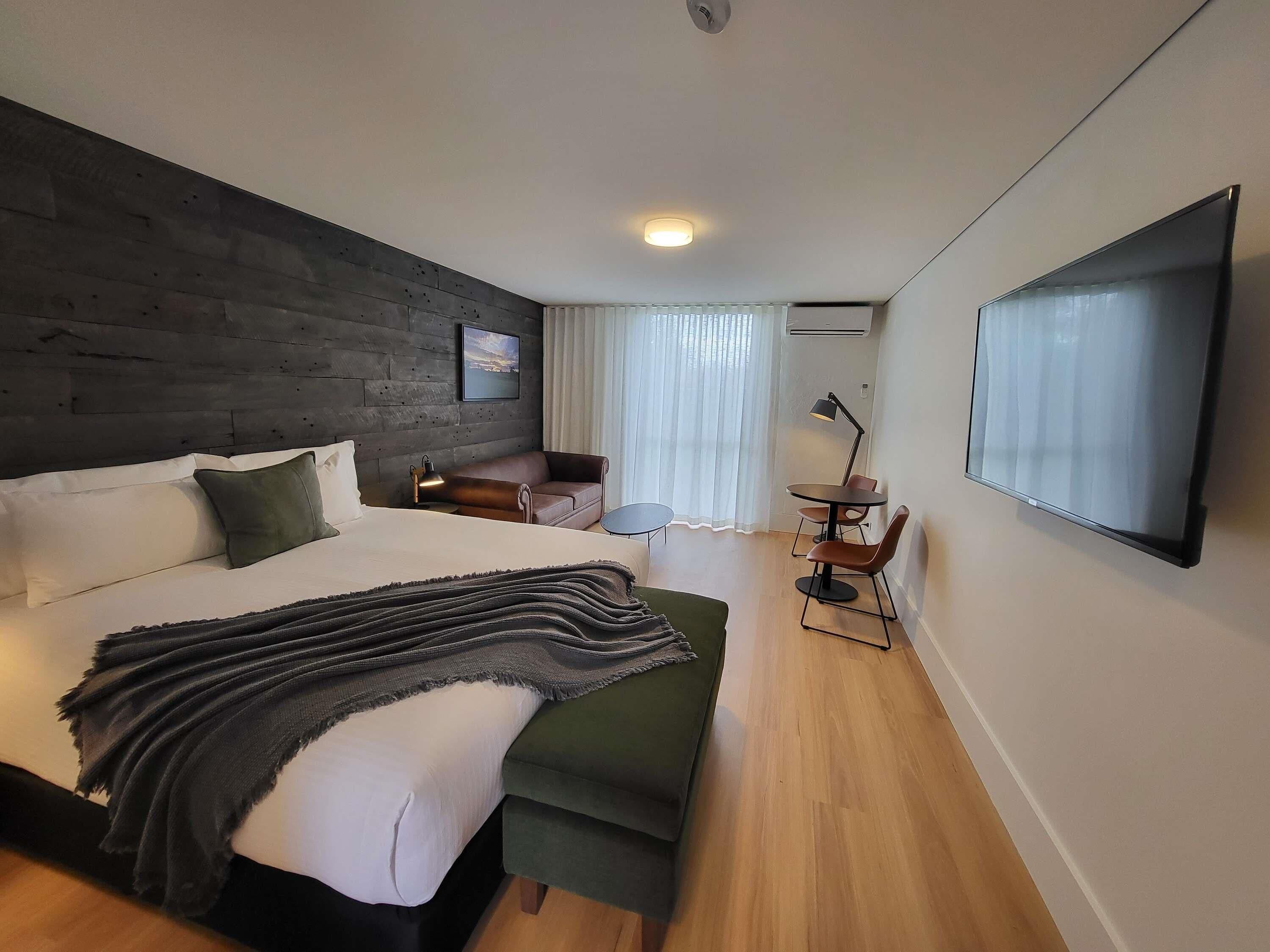 Una habitación de hotel moderna con una cama grande, un sofá verde, una mesita y un televisor montado en la pared.