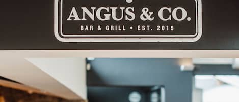 A restaurant interior with a logo for 'Angus & Co.' and a bull emblem. There are wooden tables and chairs, a stone wall, and a bar area.