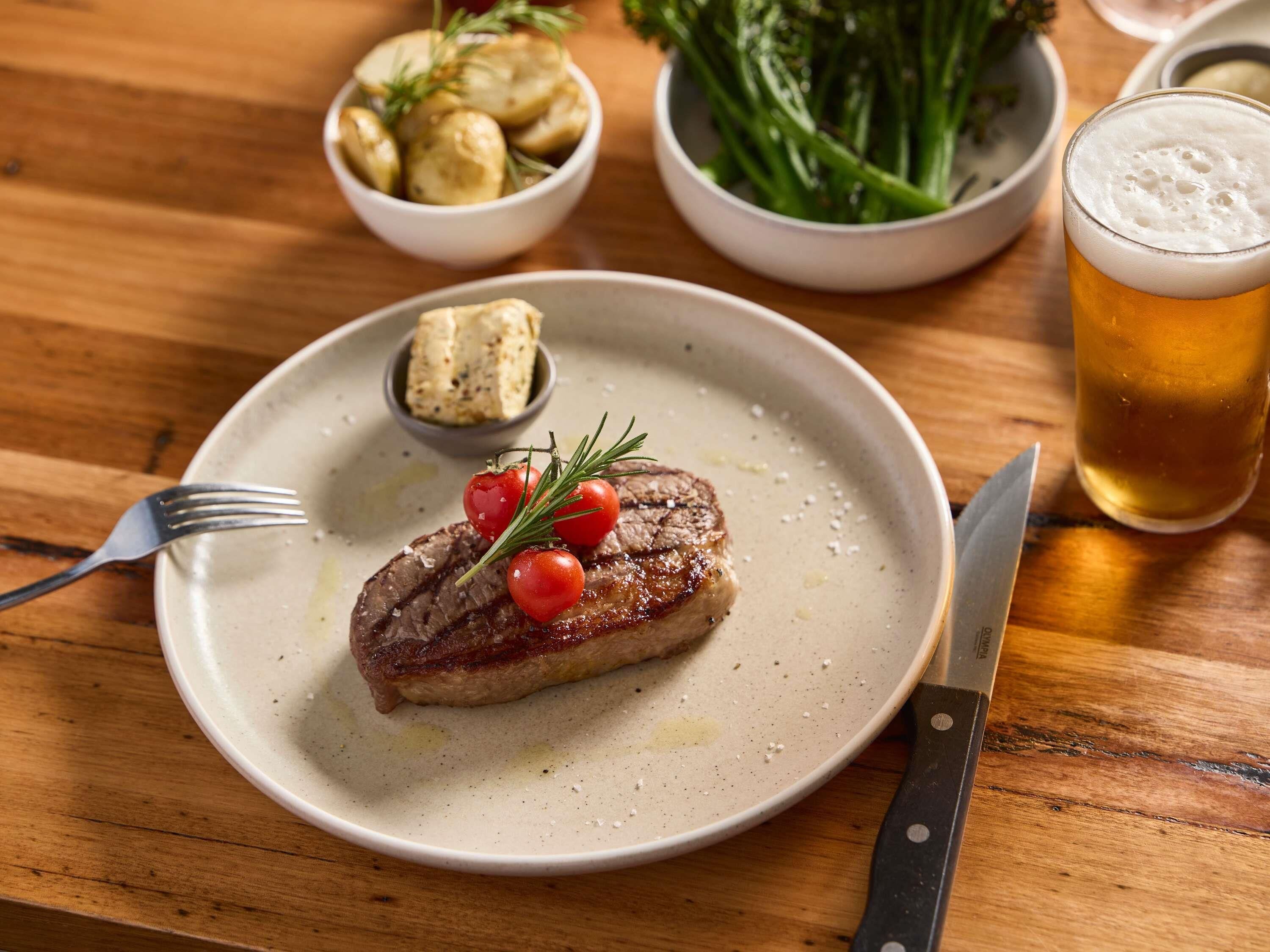 Un plato con un bistec, adornado con tomates cherry y romero, acompañado de un tenedor y cuchillo sobre una mesa de madera.