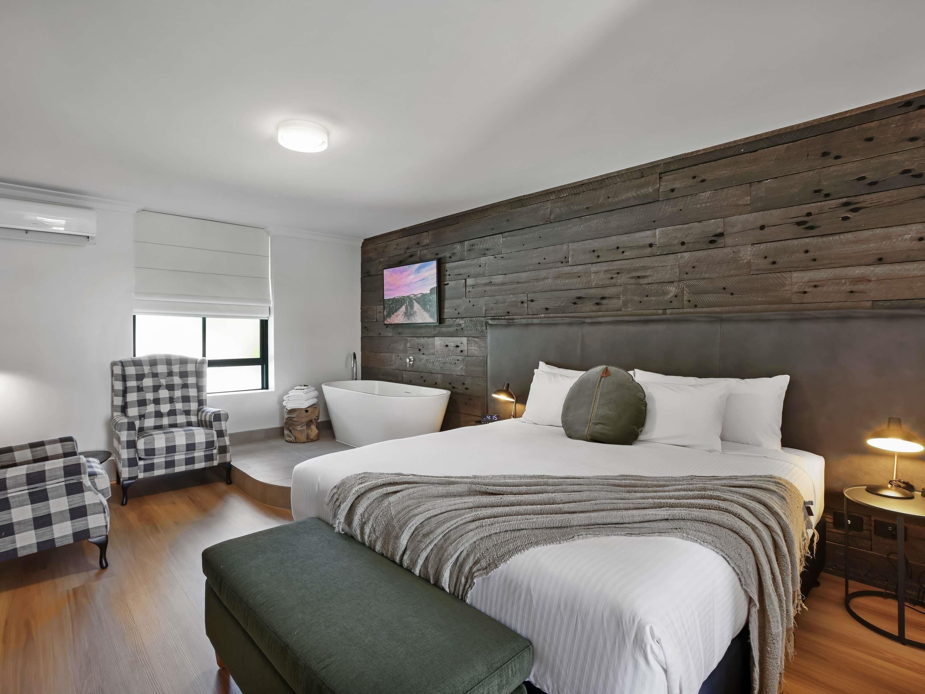 Un dormitorio moderno con una cama grande, cabecera de madera, un sillón a cuadros, una mesita auxiliar pequeña y un banco.