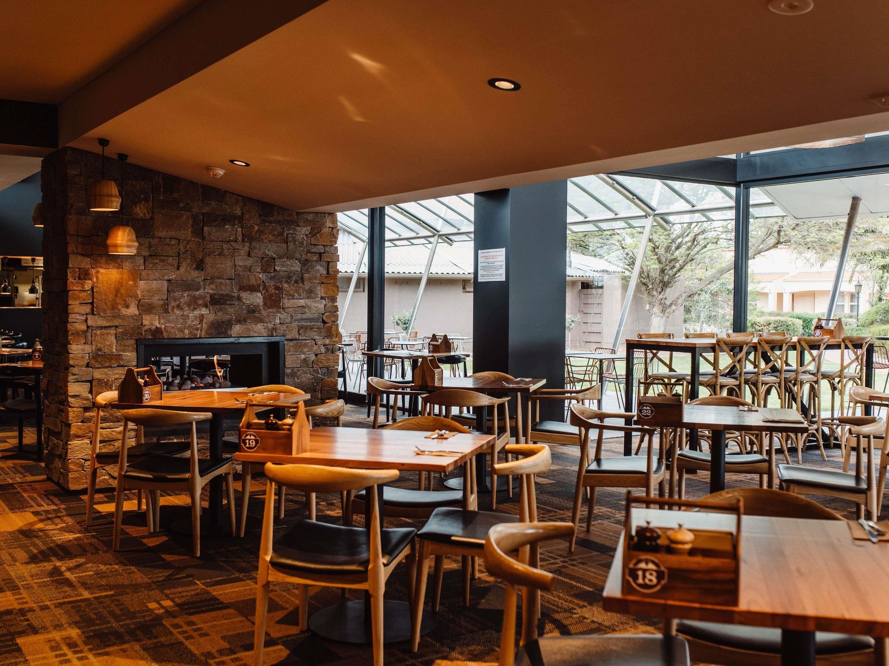 A restaurant with a stone wall, wooden tables and chairs, and large windows offering a view of the outdoors.