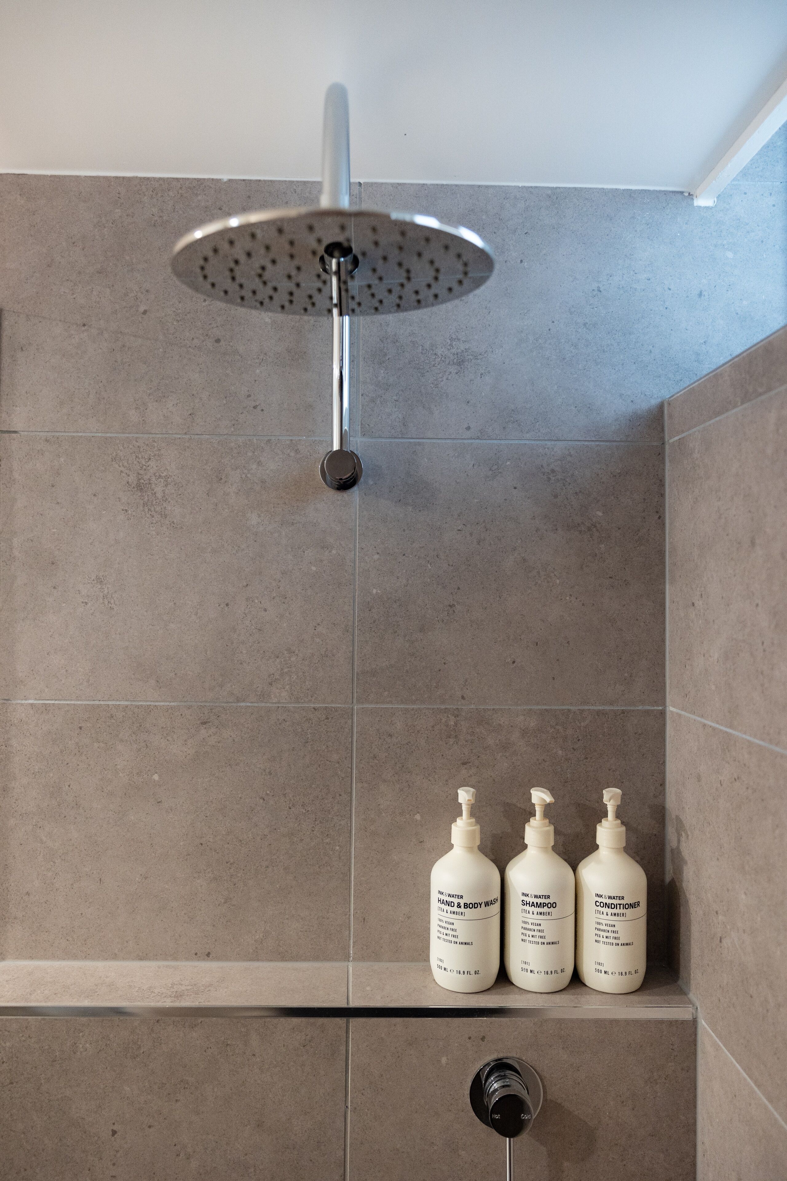 A shower area with a rain showerhead, three bottles of shampoo, and a hand and body wash dispenser.