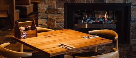 A cozy dining area with a wooden table and chairs, a stone wall, and a fireplace with a fire.