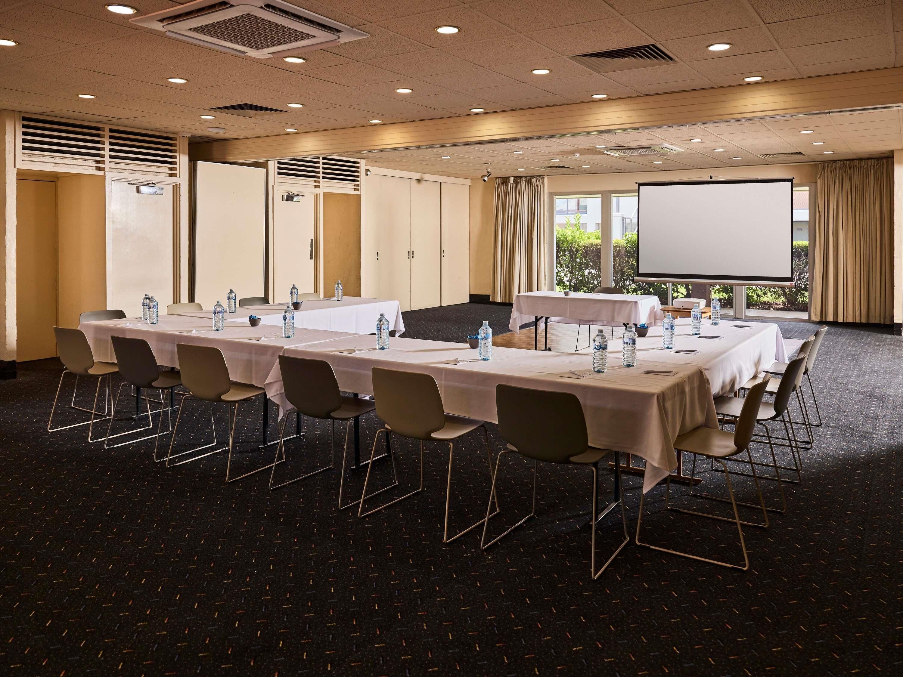 Un salón de conferencias con mesas largas, sillas y una pantalla para proyector.