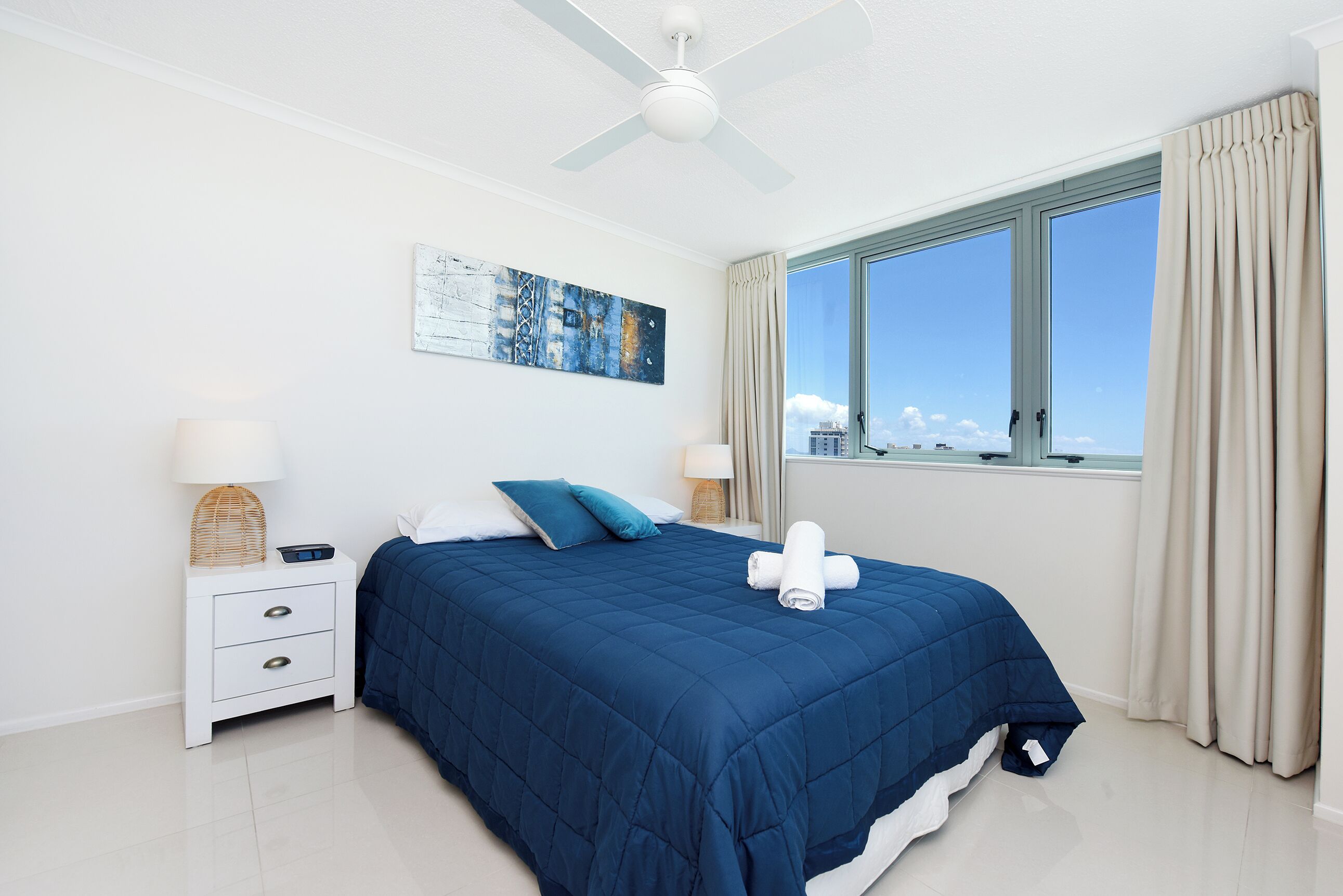 Une chambre avec un grand lit, un ventilateur de plafond, une fenêtre avec des rideaux, une table de chevet avec une lampe et un tableau au mur.