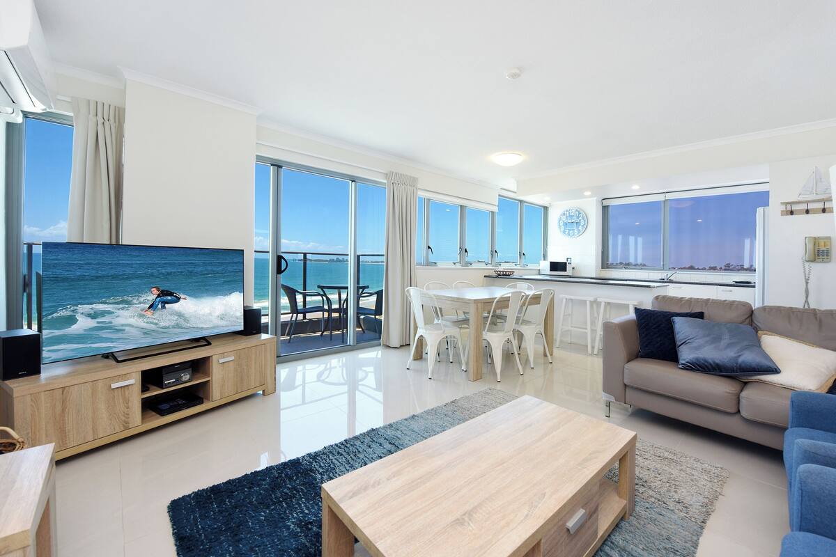 A modern living room with a large flat-screen TV, a wooden coffee table, a sofa, and a dining area with chairs.