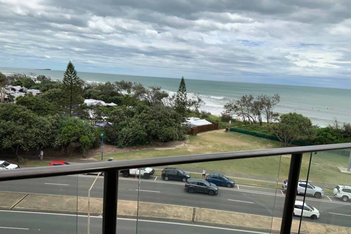 A coastal view with a road, parked cars, and a beach in the background.