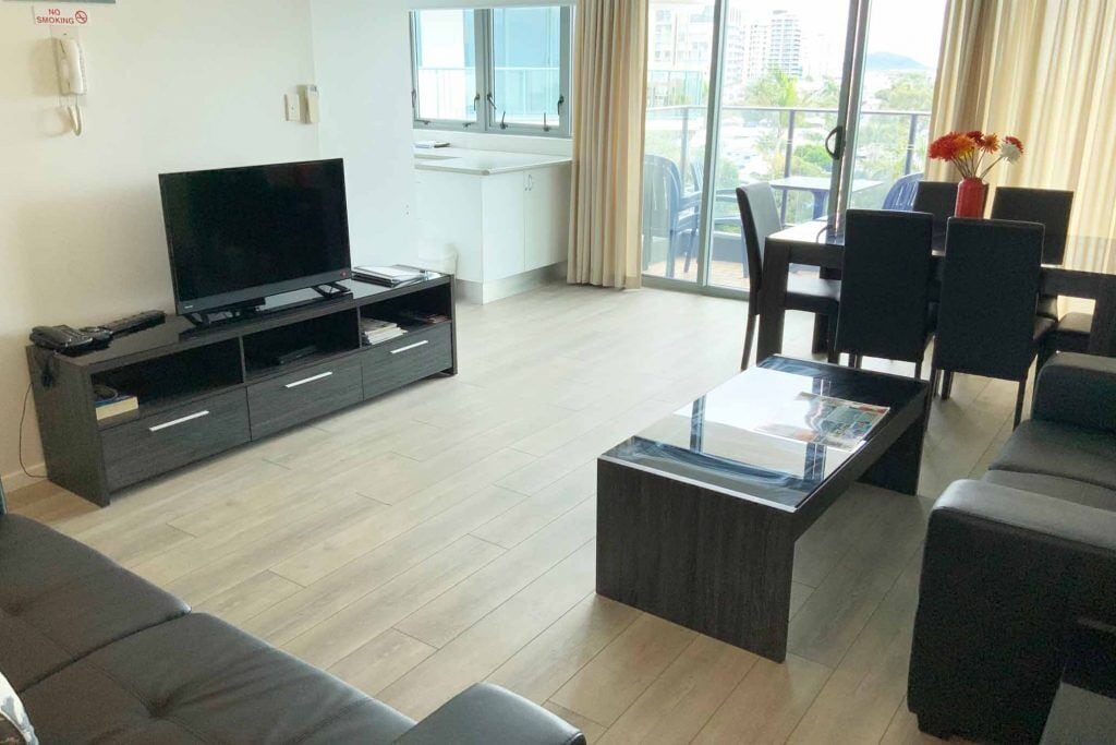 Een moderne woonkamer met een flatscreen televisie, donker meubilair, een glazen salontafel en uitzicht op de natuur door grote ramen.