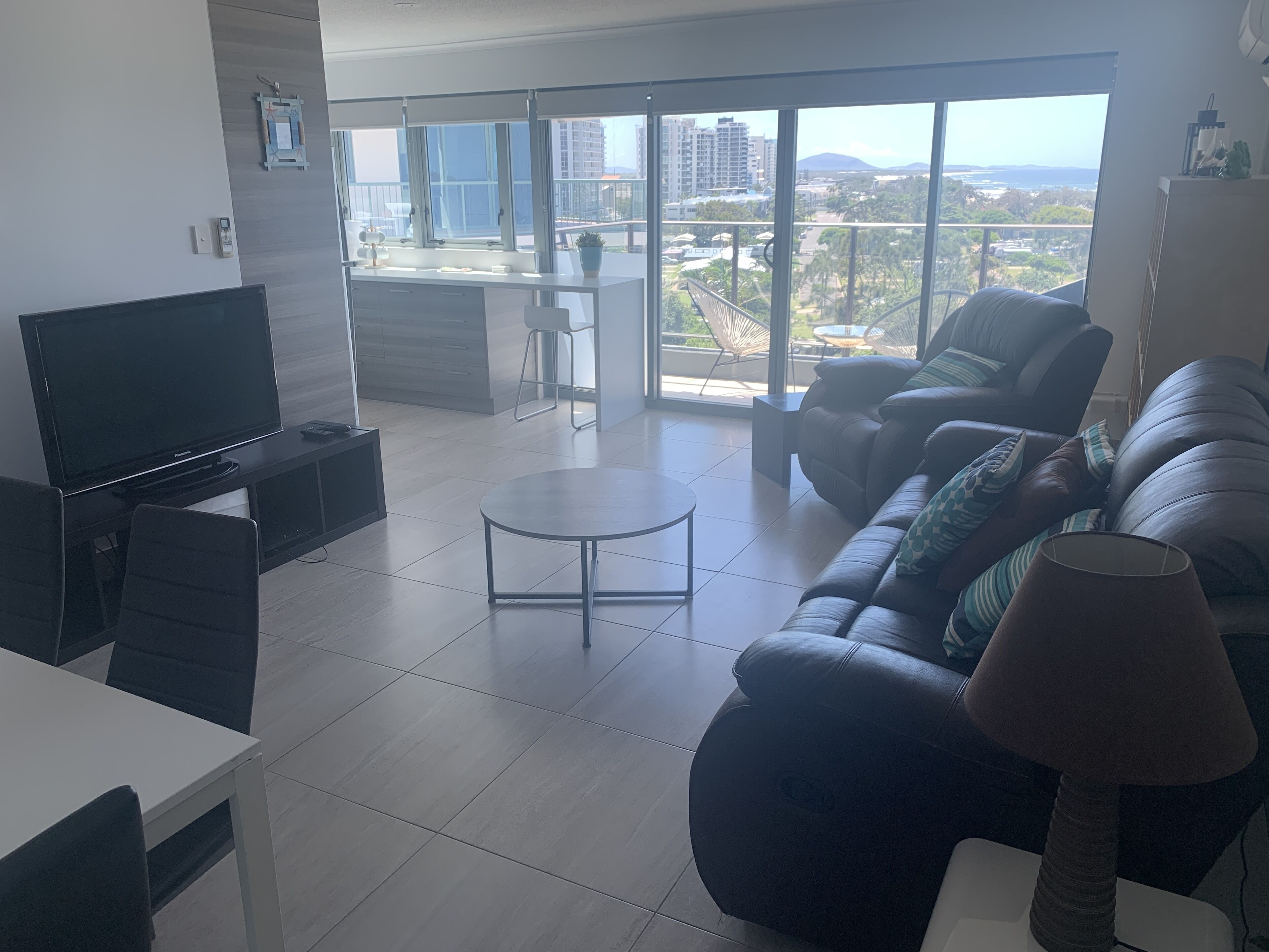 A modern living room with a flat-screen TV, a sofa, a coffee table, and a view of a balcony with a cityscape and mountains in the distance.