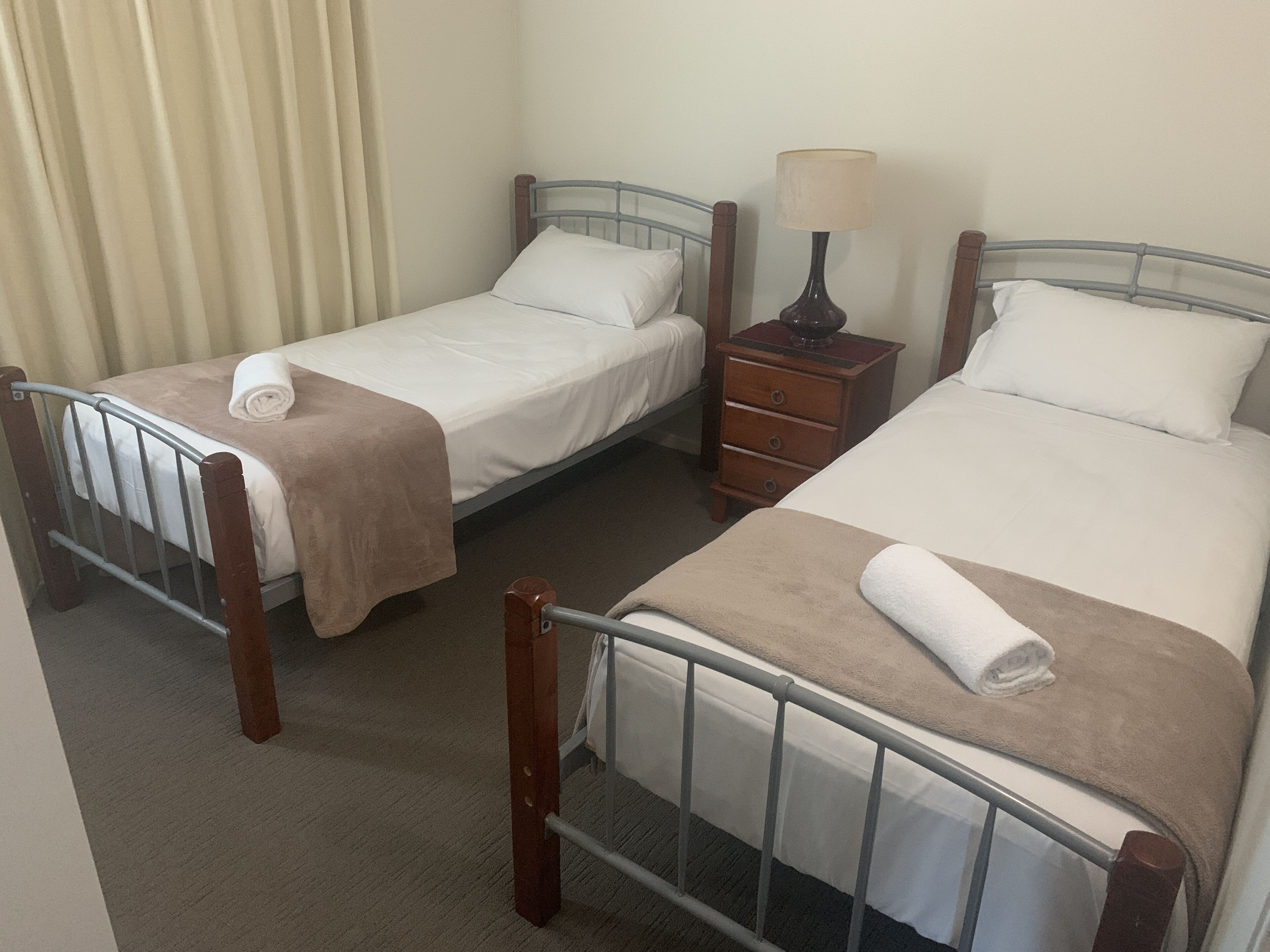 A hotel room with two single beds, a nightstand with a lamp, and a window with curtains.