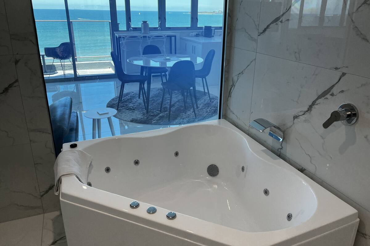 A modern bathroom with a large bathtub, a marble-tiled wall, and a view of the ocean through large windows.