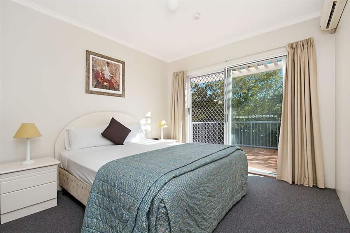 A bedroom with a bed, a nightstand, a lamp, a framed picture, and a sliding glass door to a balcony.