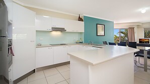 A modern kitchen with white cabinets, a central island, and a dining area with a TV. - Blue Waters Apartments (Labrador)