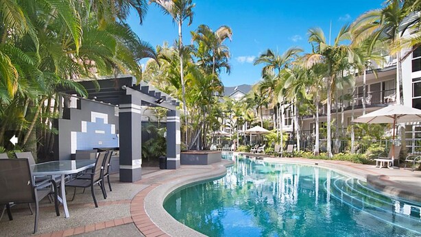 A pool area with a glass table and chairs, surrounded by palm trees and a building with balconies. - Blue Waters Apartments (Labrador)