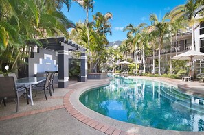 A pool area with a glass table and chairs, surrounded by palm trees and a building with balconies. - Blue Waters Apartments (Labrador)