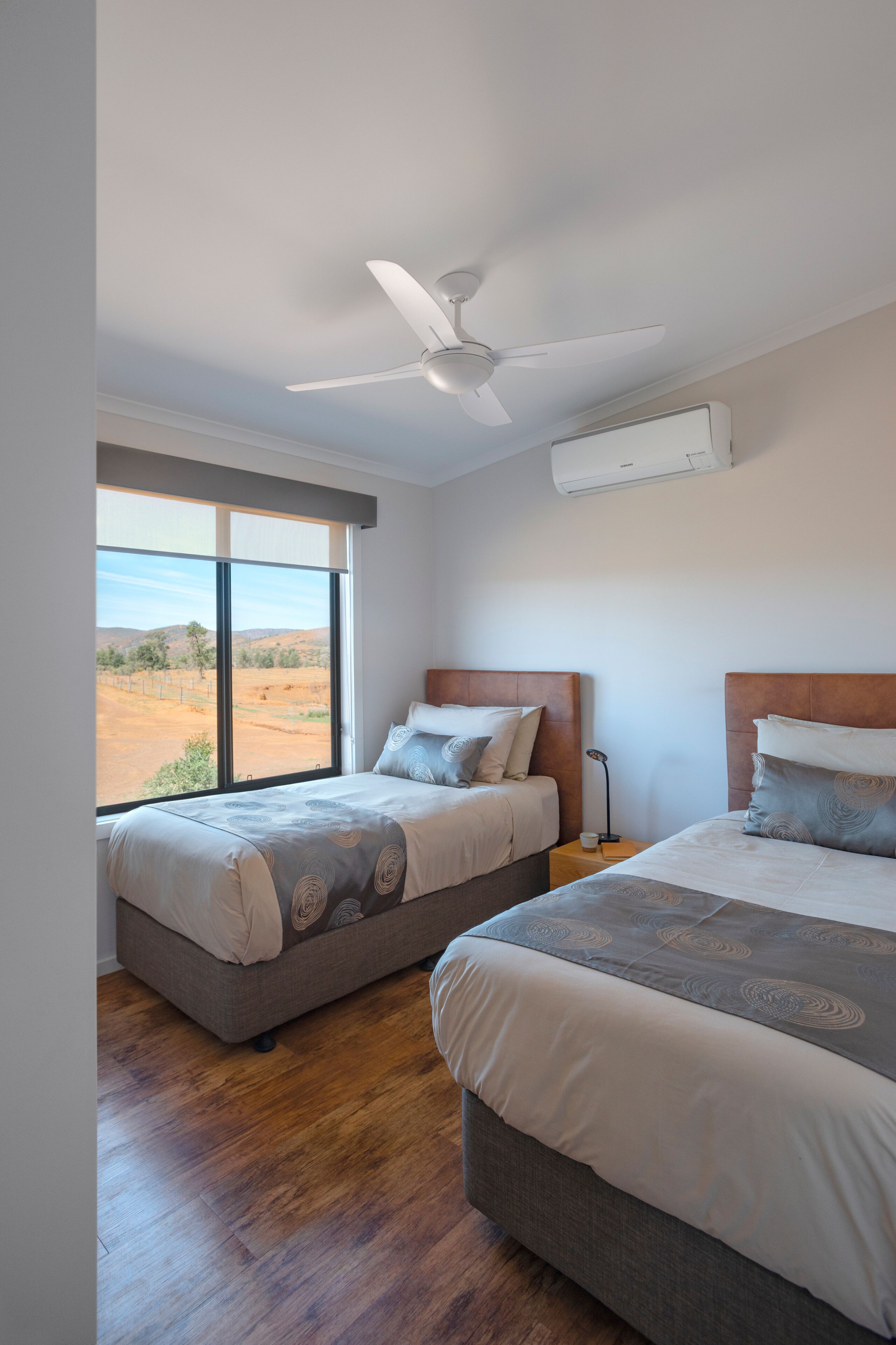 Quarto com duas camas, um ventilador de teto e uma janela grande com vista para o campo.